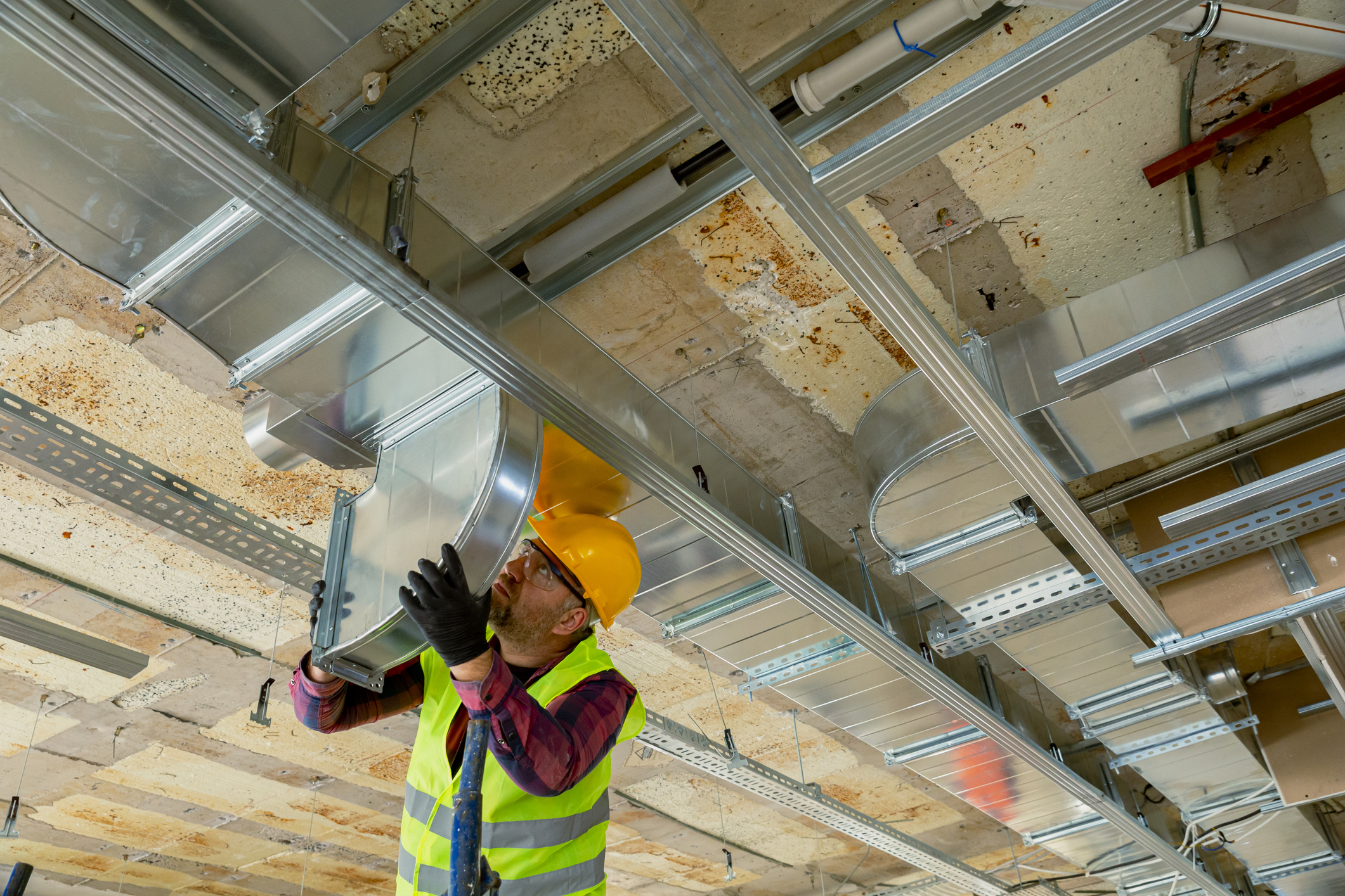 Worker in hard hat installing ductwork overhead on a construction site