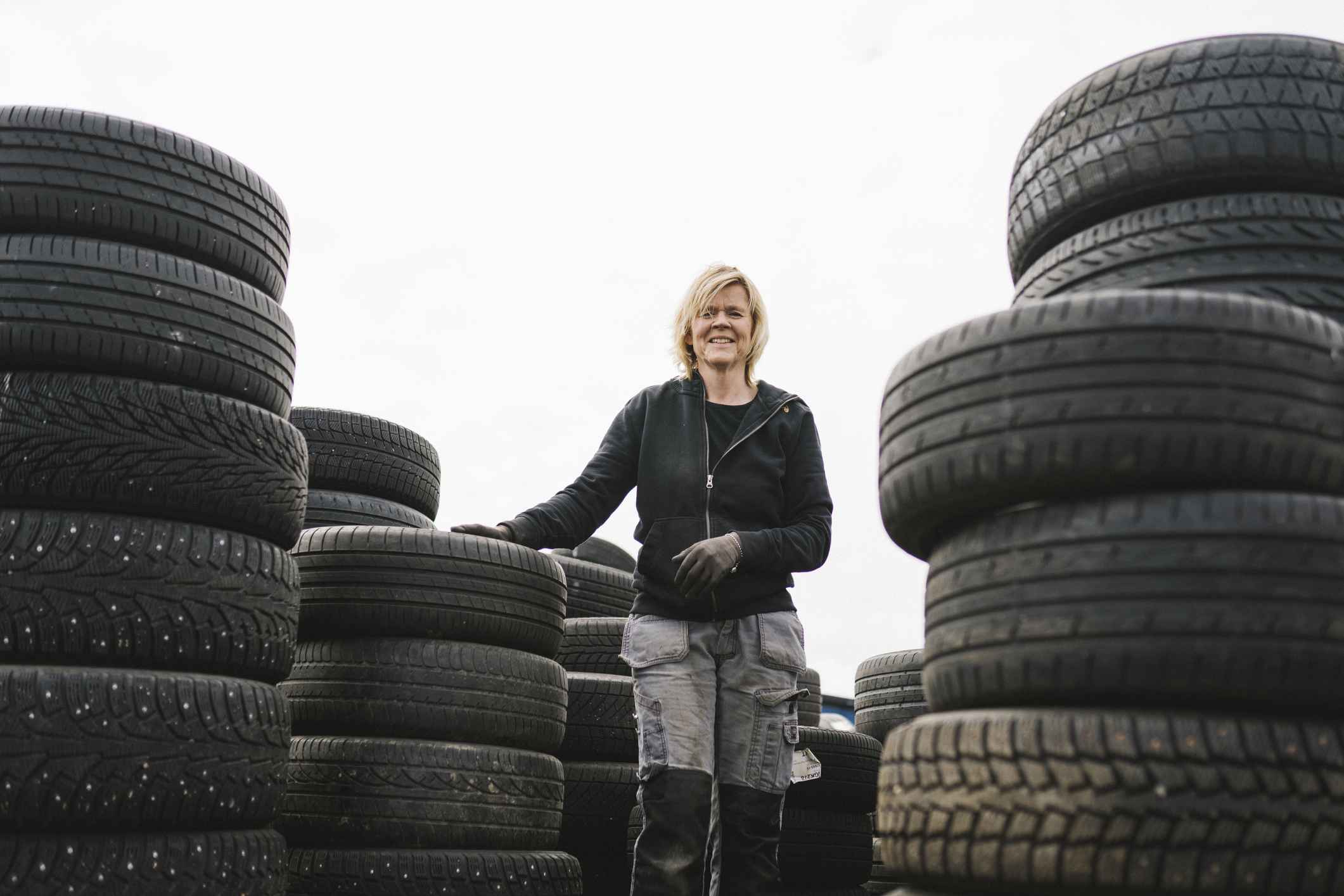 Woman standing between stacks of tires at a workshop, smiling, dressed in work attire