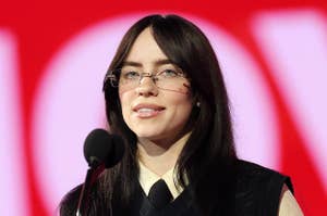 Billie Eilish speaking at a podium, wearing a black outfit and patterned glasses