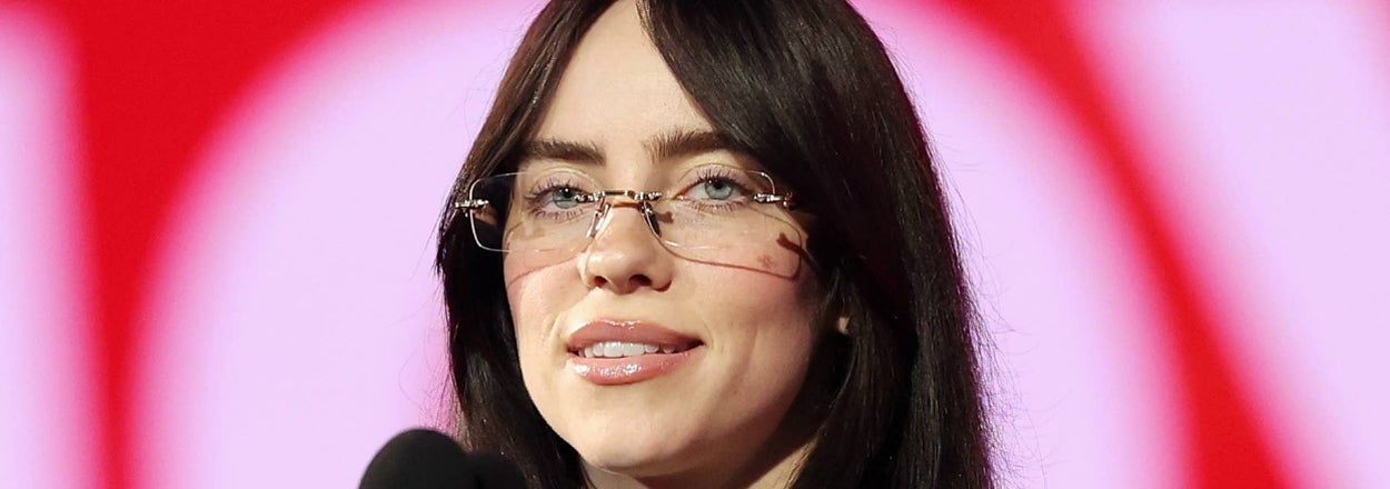 Billie Eilish speaking at a podium, wearing a black outfit and patterned glasses