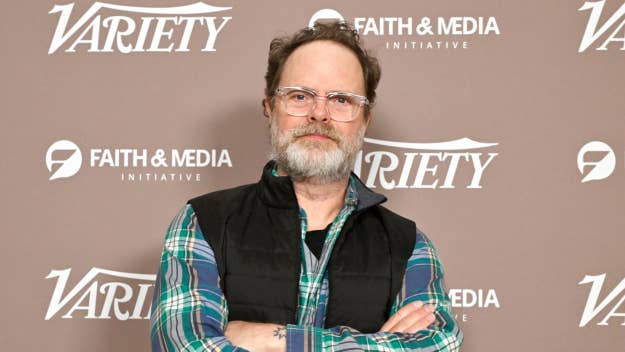 Rainn Wilson poses in front of a backdrop with crossed arms, wearing glasses and a vest over a long-sleeve shirt