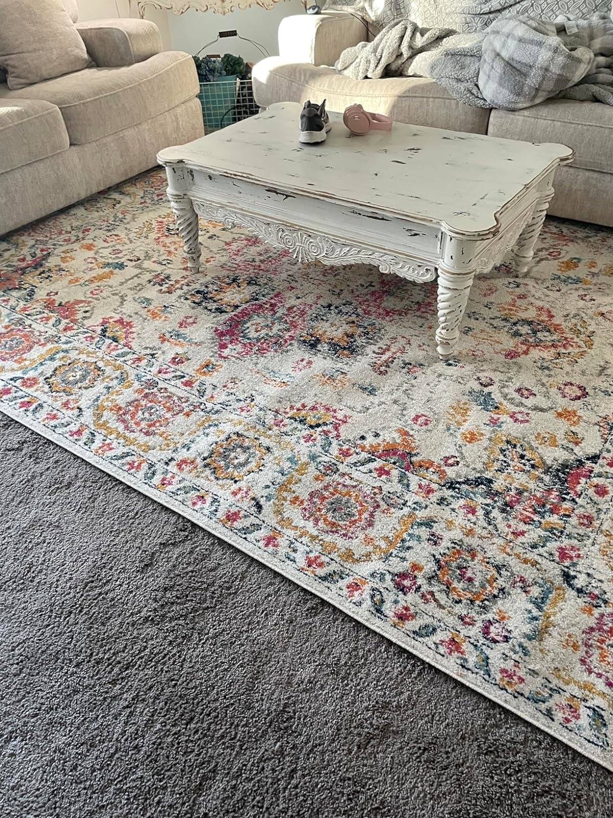 A distressed white coffee table on a floral rug in a living room, with a sofa and blanket visible