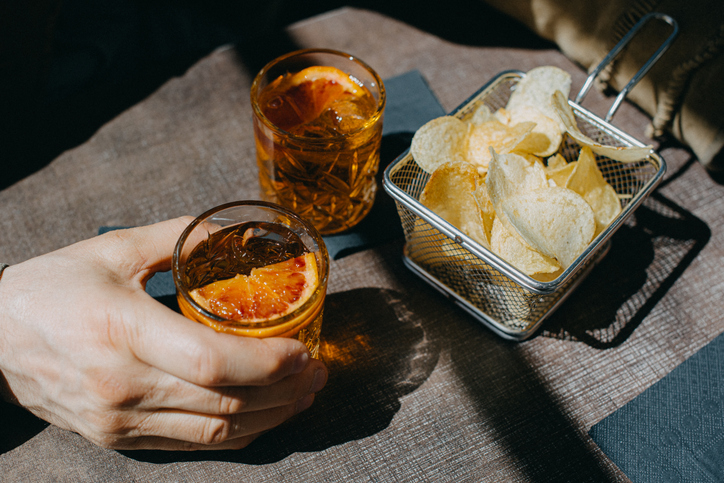 Hand holding a glass with a citrus garnish beside a basket of chips in sunlight