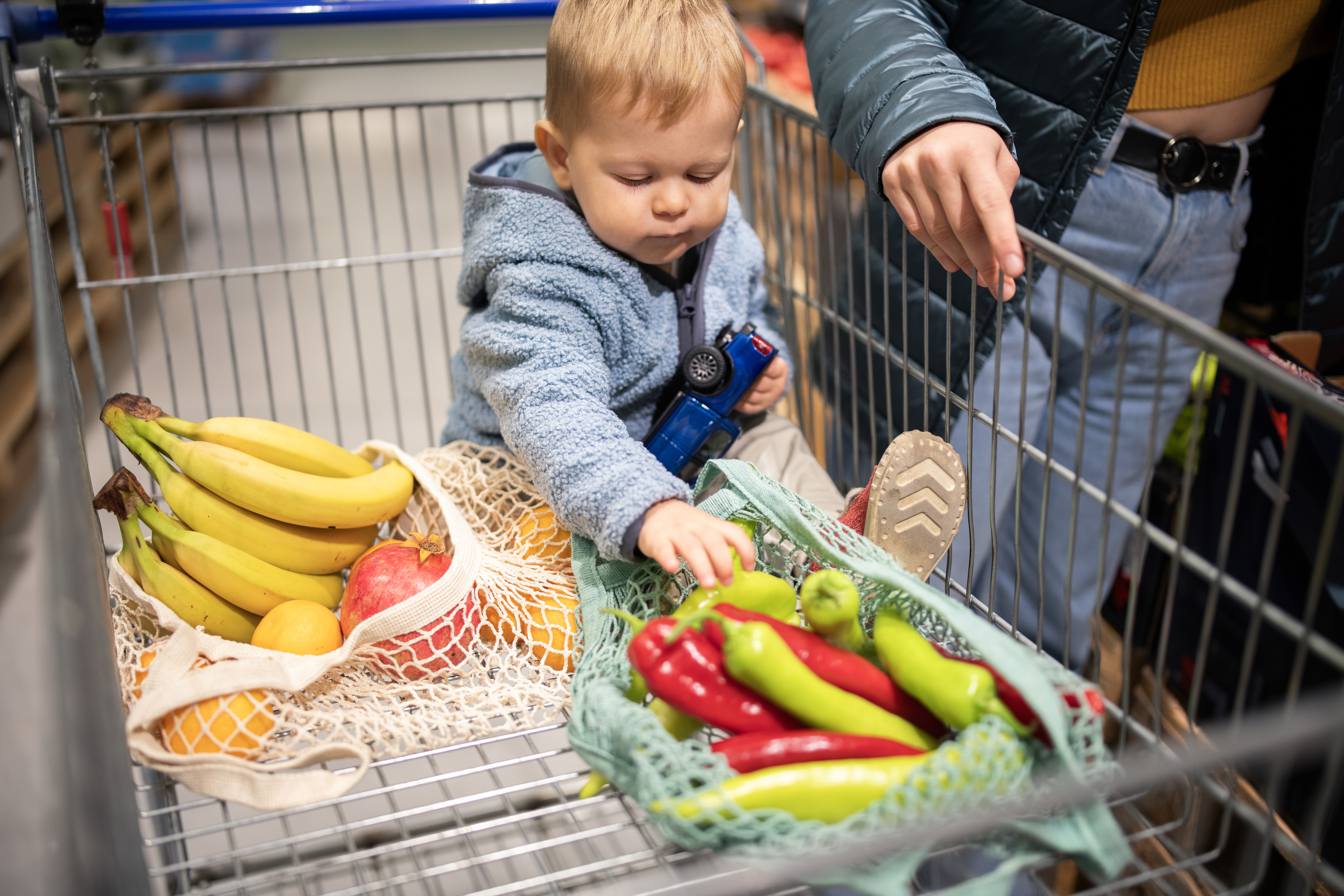 Toddler sitting in a shopping cart with fruits and vegetables, under the supervision of an adult