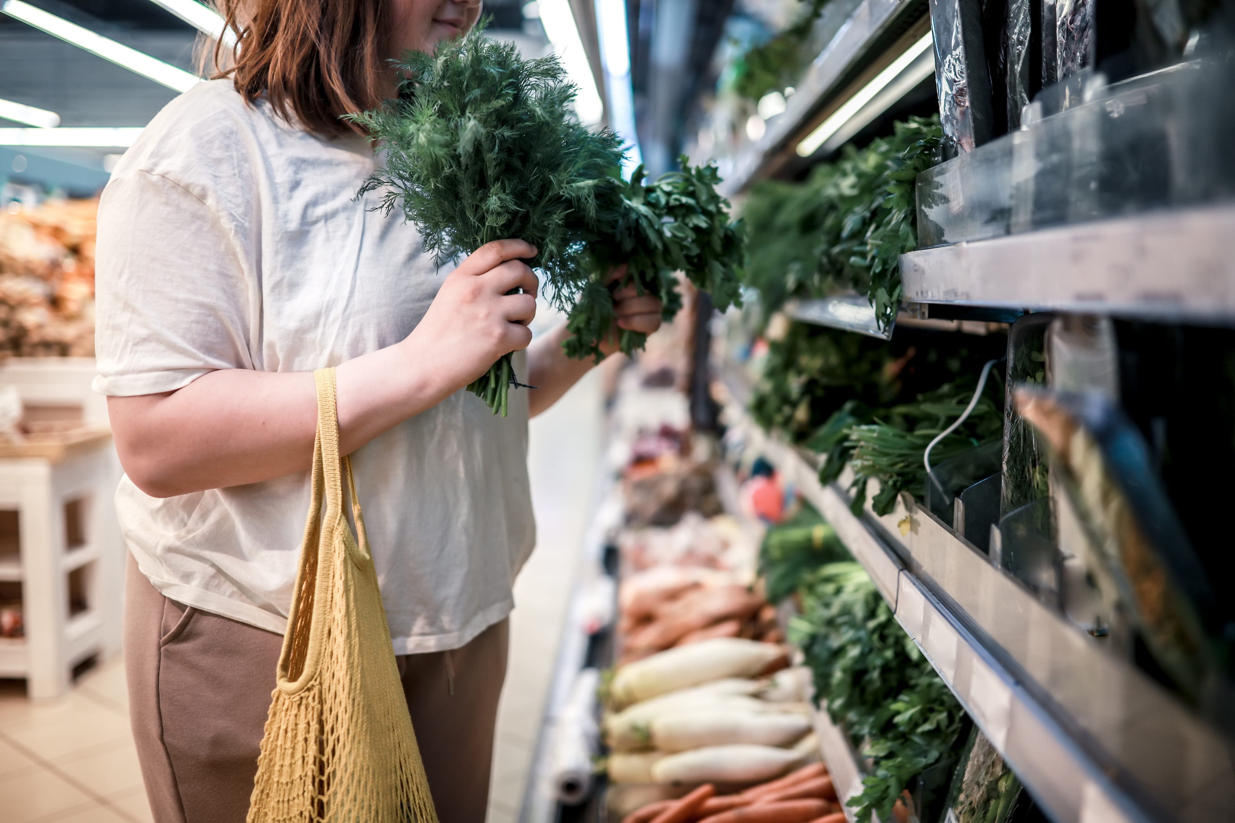 Person shopping for vegetables in a grocery store, holding dill and a reusable bag