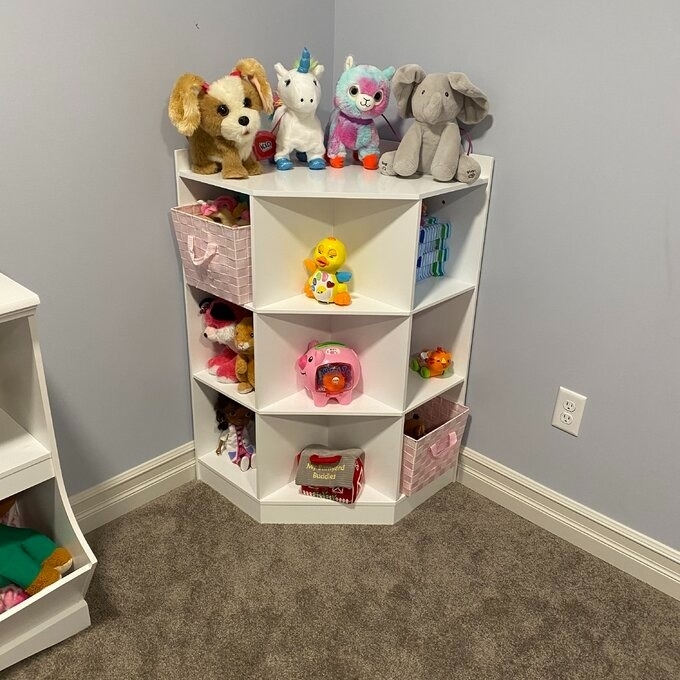 A white corner shelf filled with various children&#x27;s toys including stuffed animals and toy vehicles in a room