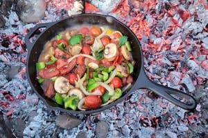 Cast-iron skillet with various vegetables cooking over an open campfire