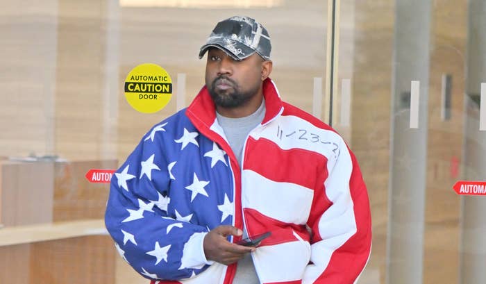 Kanye West in an American flag jacket and black boots walking past a glass door