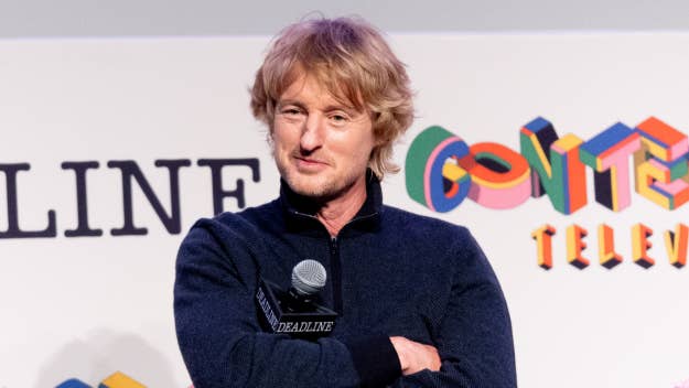 Owen Wilson at an event with microphone, casual attire, speaking onstage with logos in the background