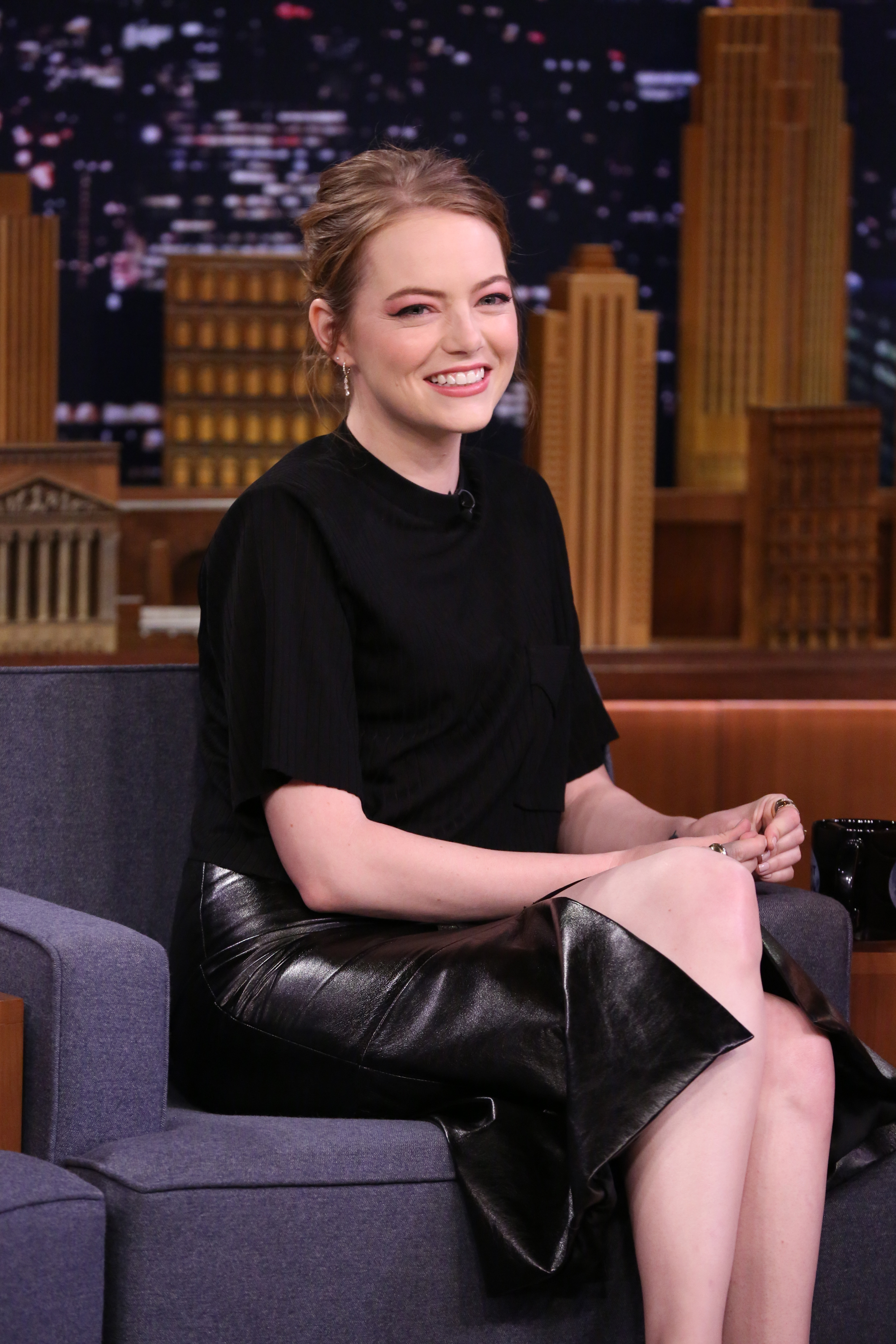 Emma Stone seated wearing a black top and leather skirt, smiling during a talk show interview