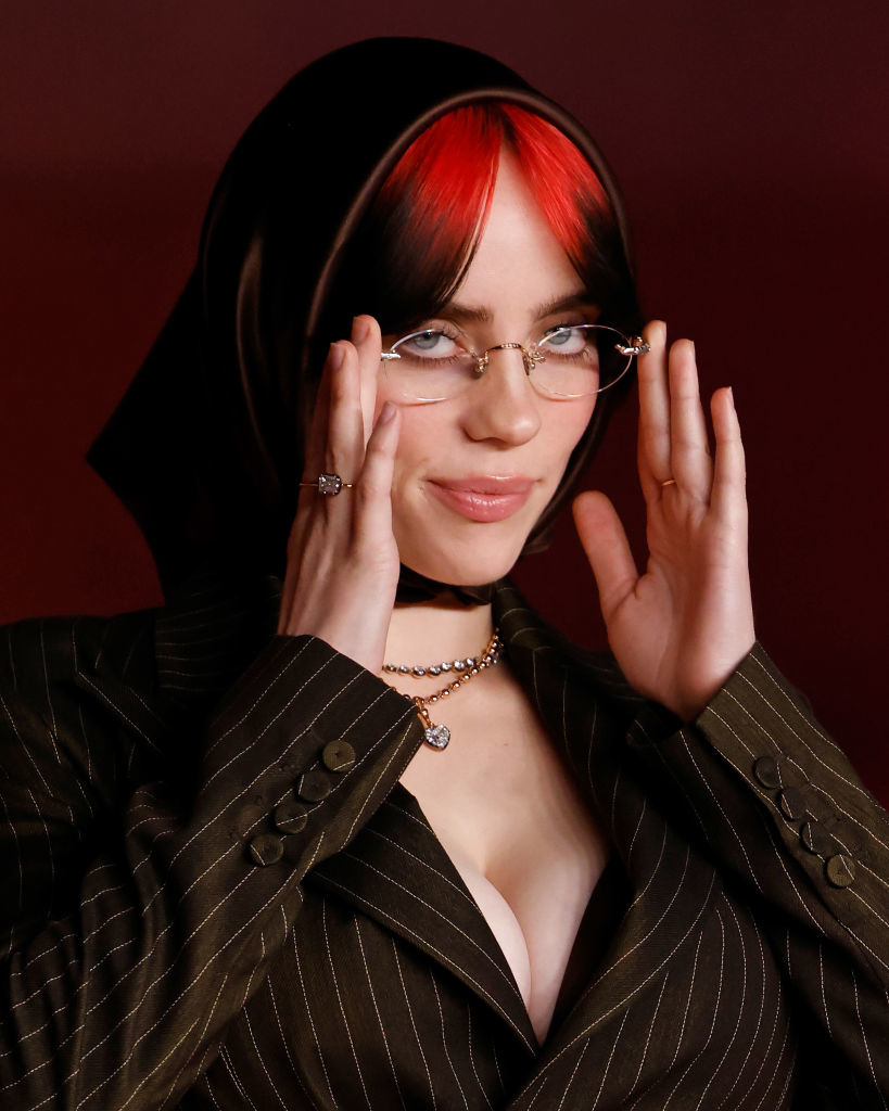 Billie Eilish in a pinstripe suit adjusts her glasses, with a red streak in her hair