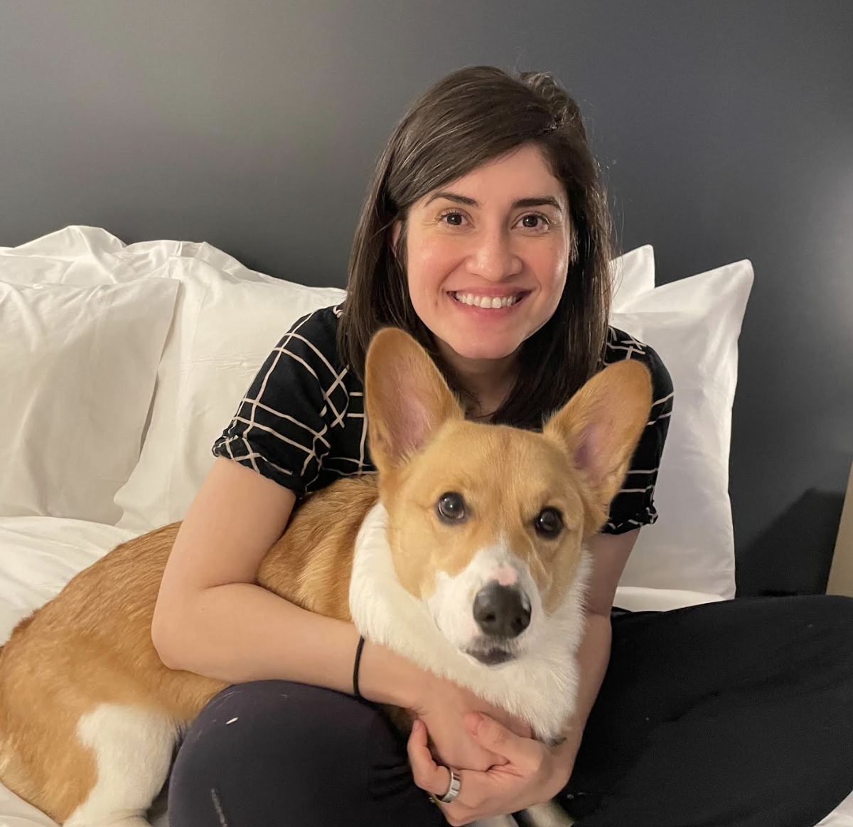 Woman smiling, sitting on a bed with a Corgi, in a cozy hotel room setting