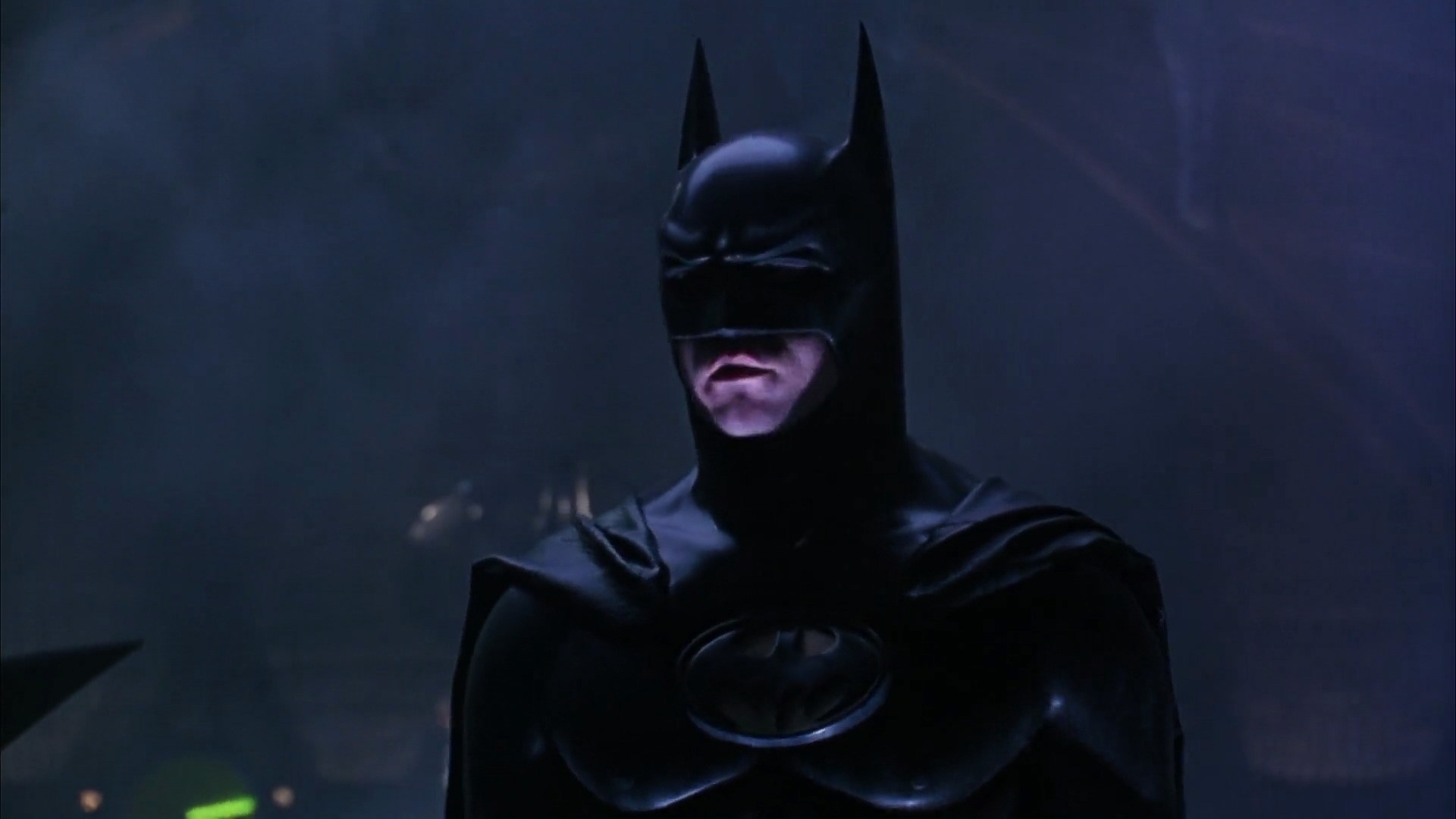 Person in a Batman costume with cowl, cape, and emblem, standing with a serious expression