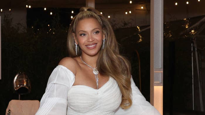 Beyoncé smiling, wearing an elegant off-shoulder white dress with sparkling earrings and a necklace