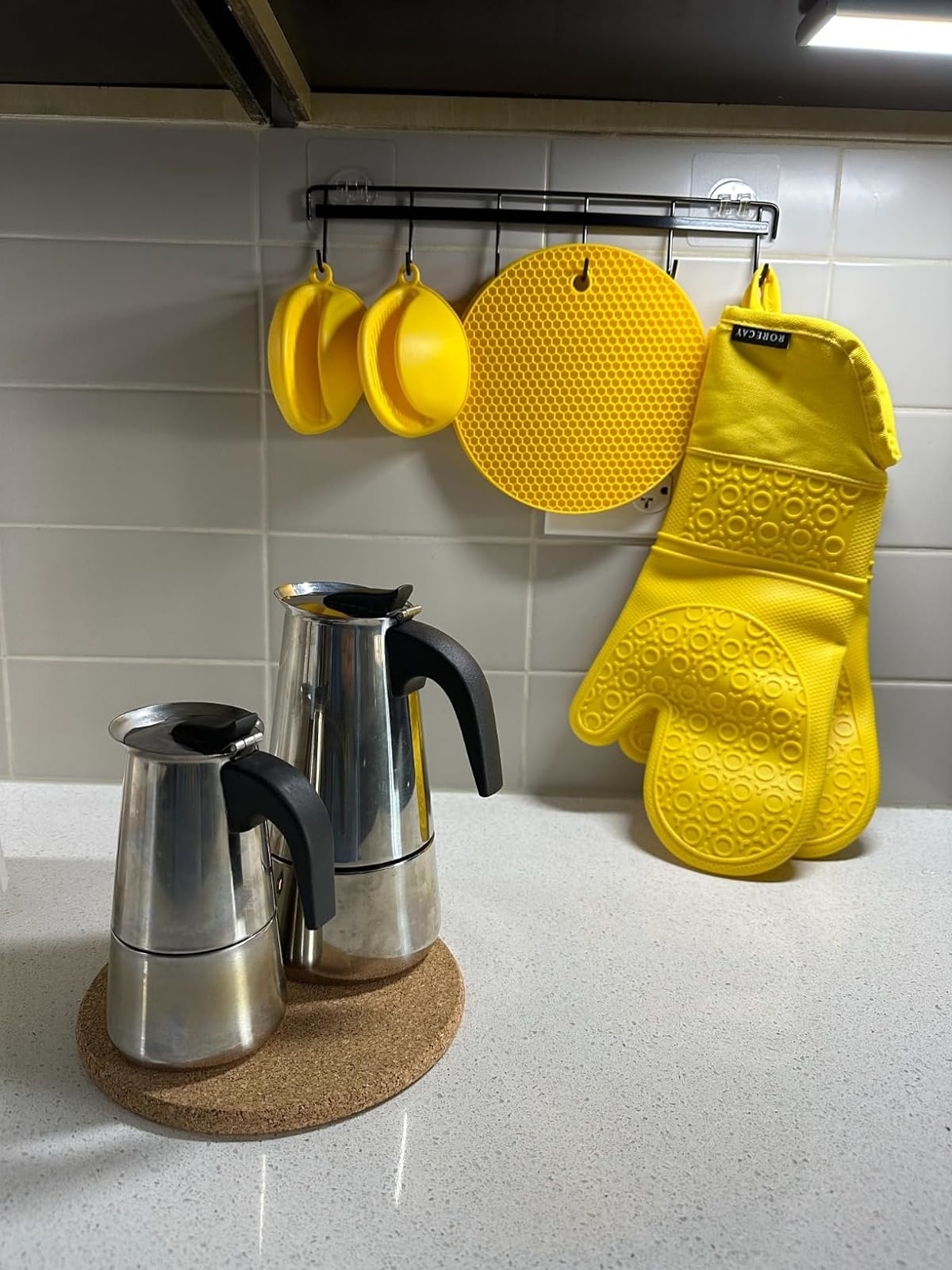 Two stainless steel coffee pots on a cork trivet, beside yellow oven mitts hanging on a kitchen rack