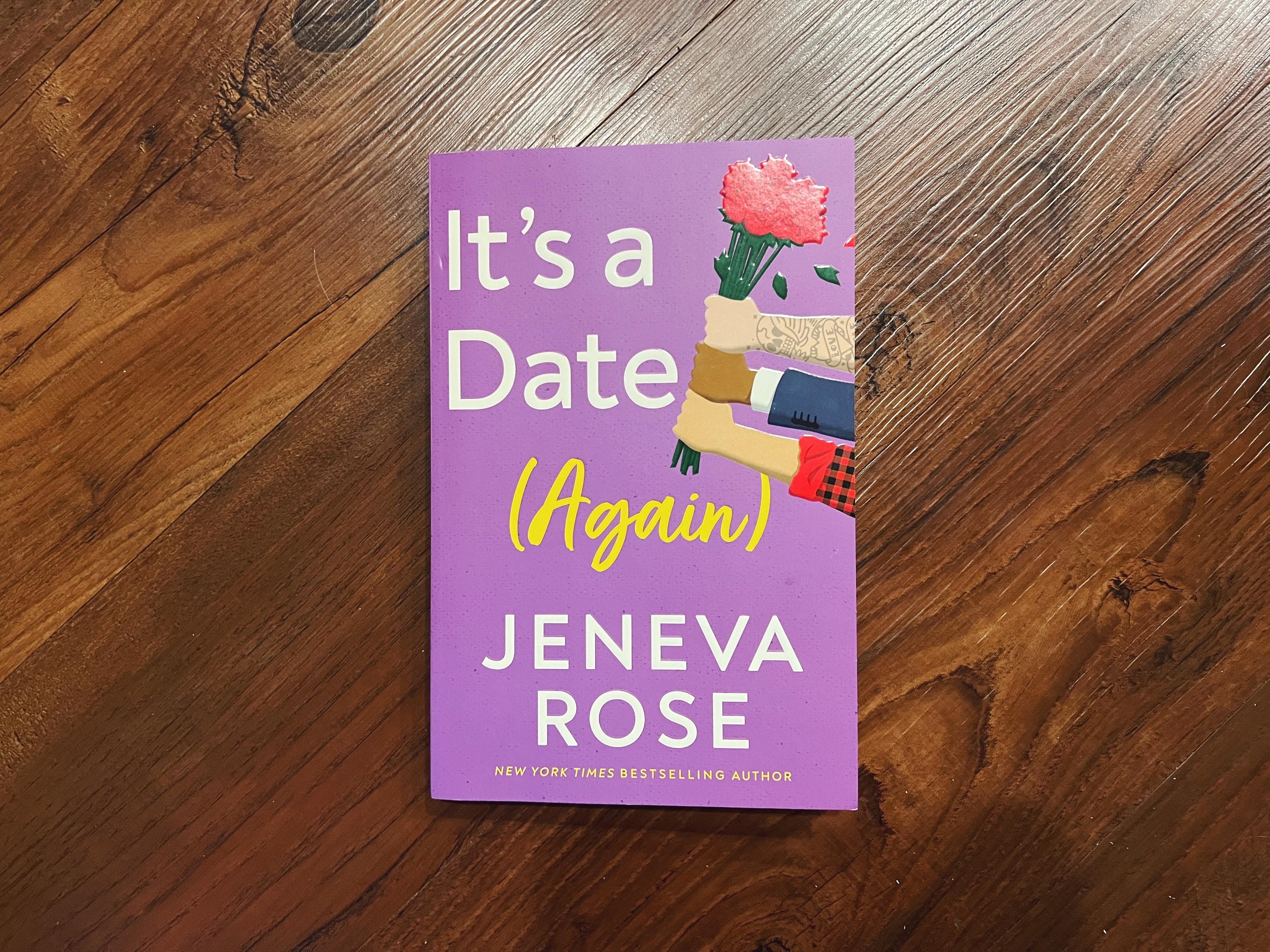 A book titled &quot;It&#x27;s a Date (Again)&quot; by Geneva Rose lays on a wooden surface