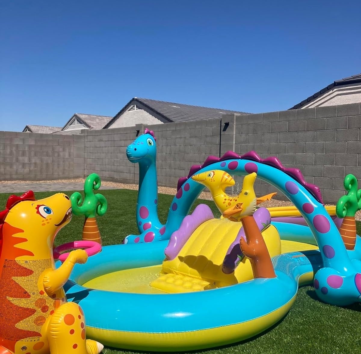 Inflatable dinosaur-themed children&#x27;s pool with sprinkler and slide in a backyard