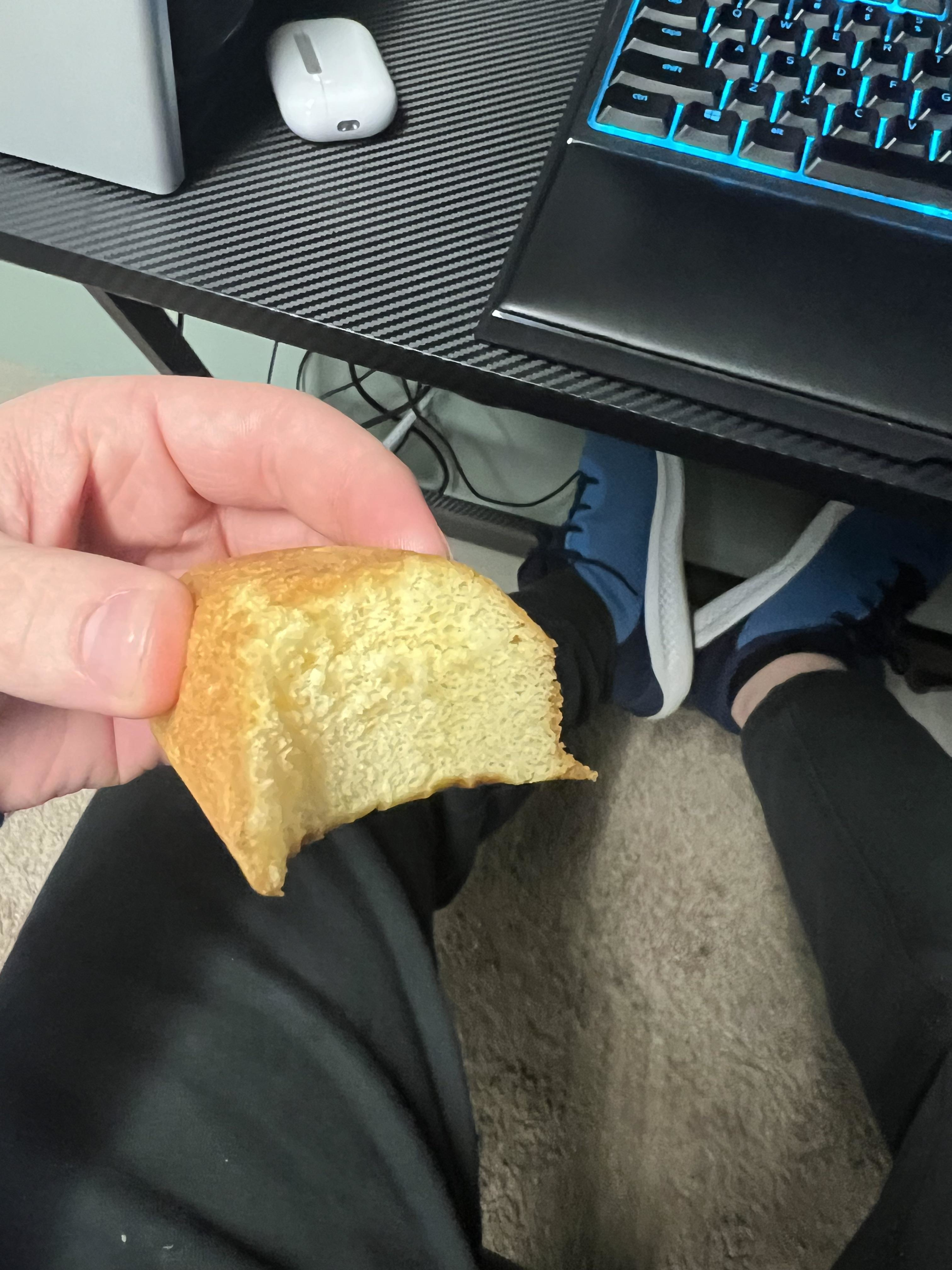 Person holding a half-eaten piece of toast with computer and legs in background