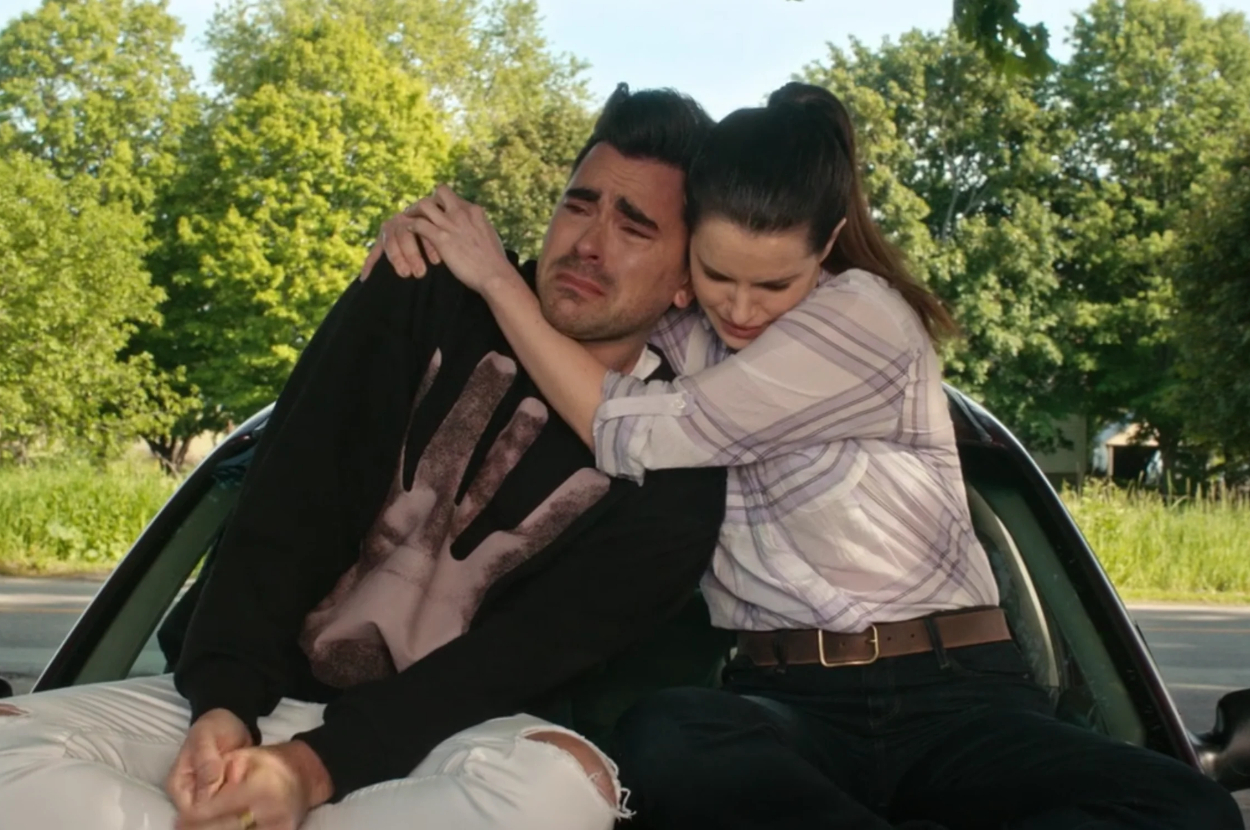 Two characters from a show express distress while embracing, sitting on a car&#x27;s trunk