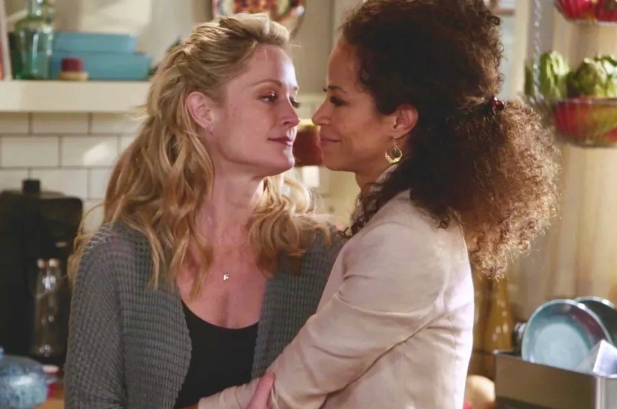 Two women embracing in a kitchen, one about to kiss the other on the cheek. They are characters in a TV show