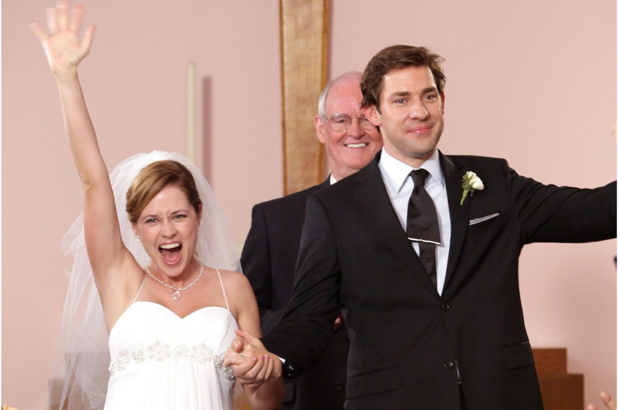 Bride in a white dress and groom in a black suit raise their hands in joy at their wedding, officiant in the background