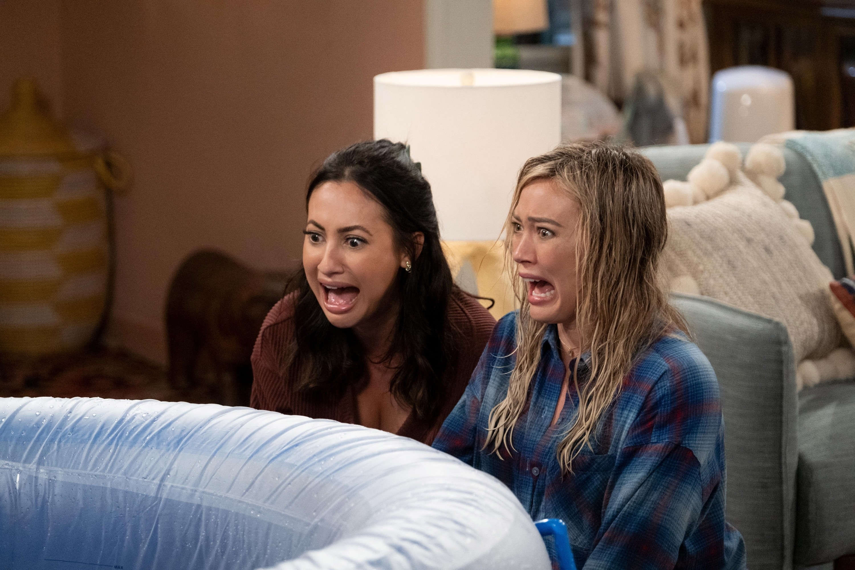 Two characters from a TV show react with shock while sitting on a couch, one in a plaid shirt