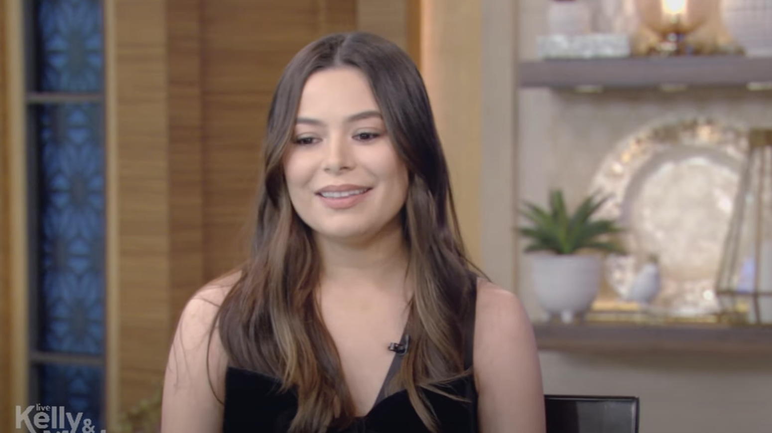 Miranda Cosgrove smiling, wearing a black top, on the set of a talk show