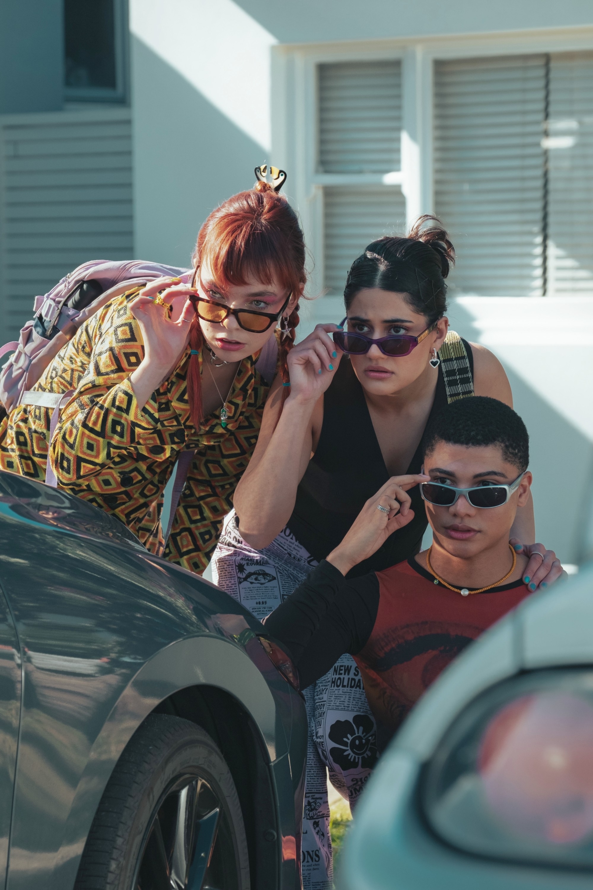 Three actors in a scene from a show, wearing stylish outfits, standing by a car with sunglasses