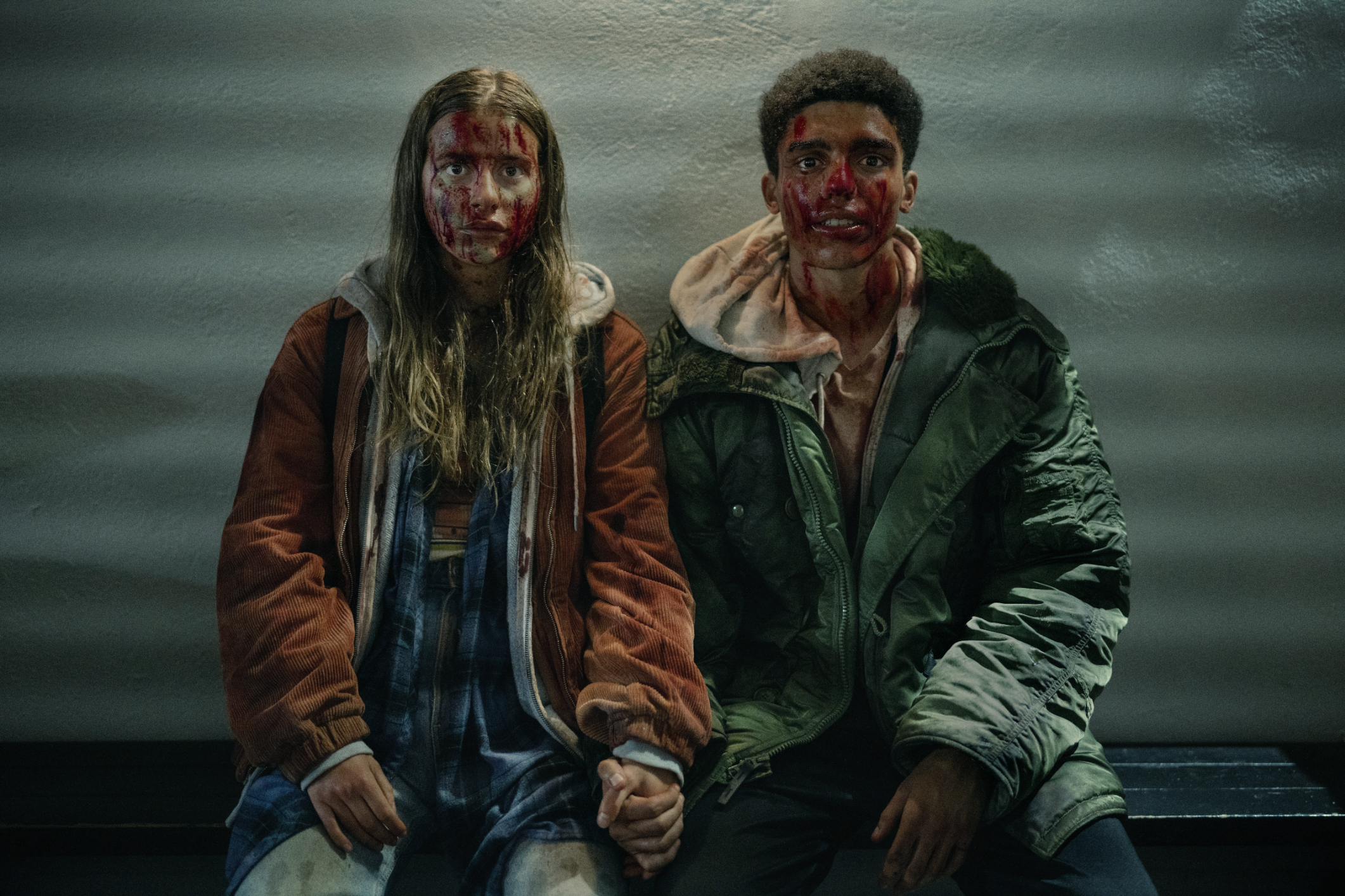 Two actors in distressed clothing, with dramatic makeup simulating injuries, sitting side by side