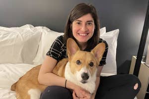 Woman smiling on a bed, hugging a corgi, in a casual setting, possibly in a hotel room