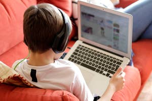 A child with headphones uses a laptop on a couch, browsing with one hand on the touchpad