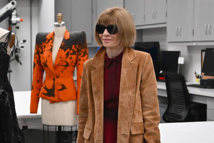 Anna Wintour in a tan coat, walking by mannequins in designer attire