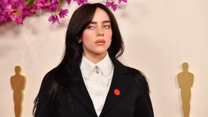 Billie Eilish at an event wearing a black blazer over a white shirt, with a red lapel pin