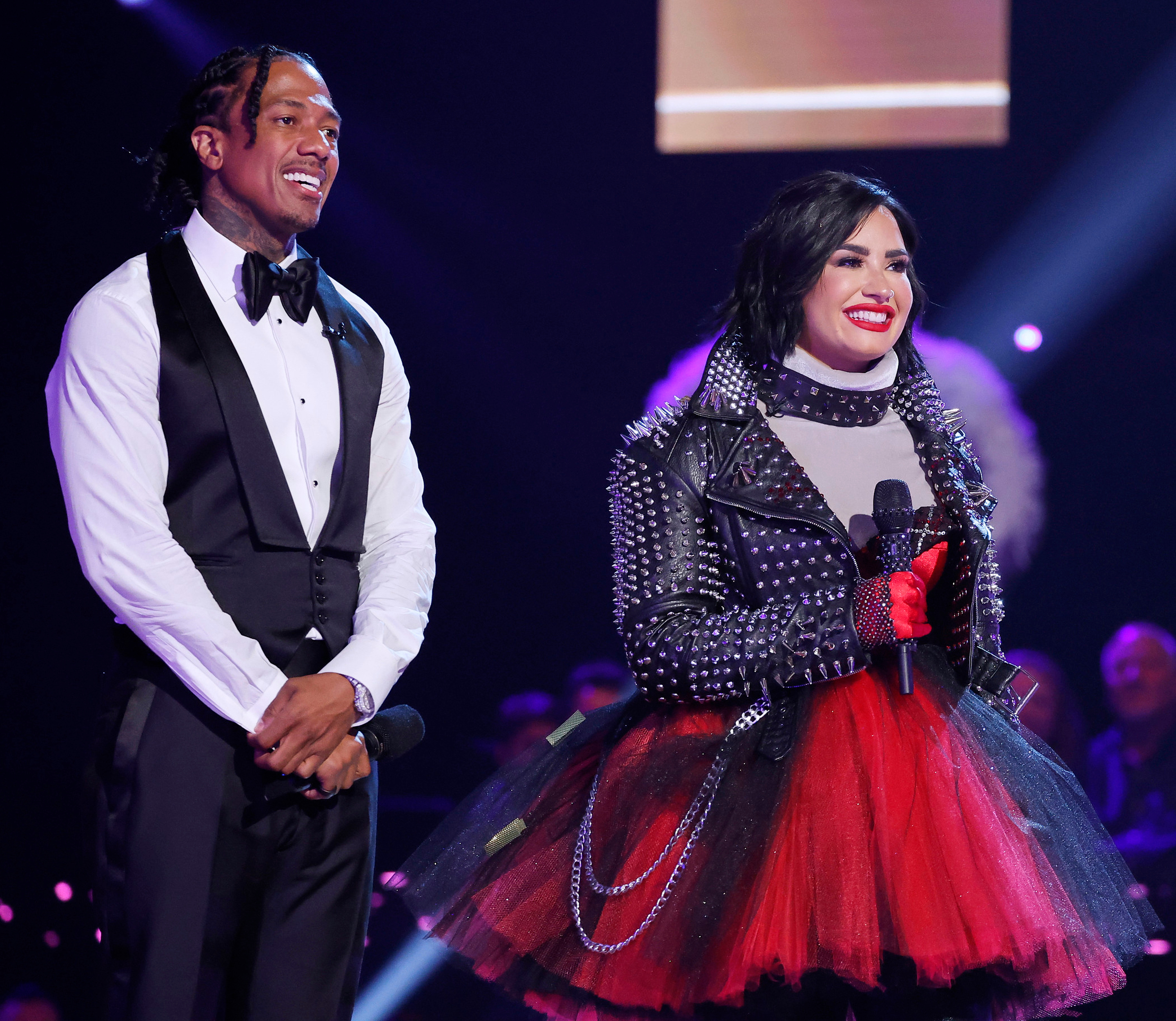 Two people on stage, one in a tuxedo, and the other in a studded jacket and tulle skirt, smiling with a microphone in hand