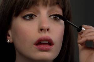 Close-up of a Anne Hathaway applying mascara to upper eyelashes, looking intently into the mirror