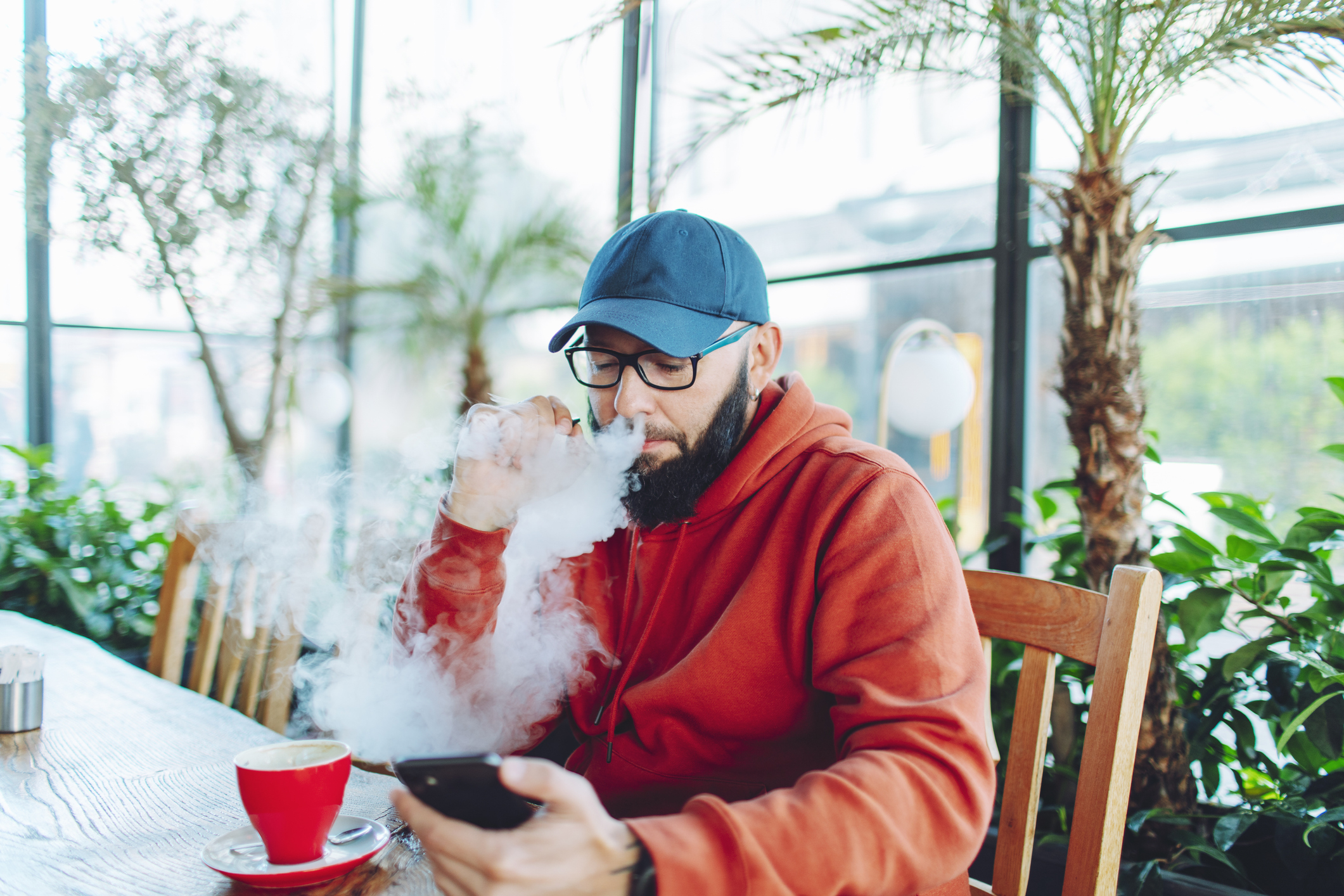 Man in a cap using a phone while exhaling vapor from an e-cigarette at a cafe table