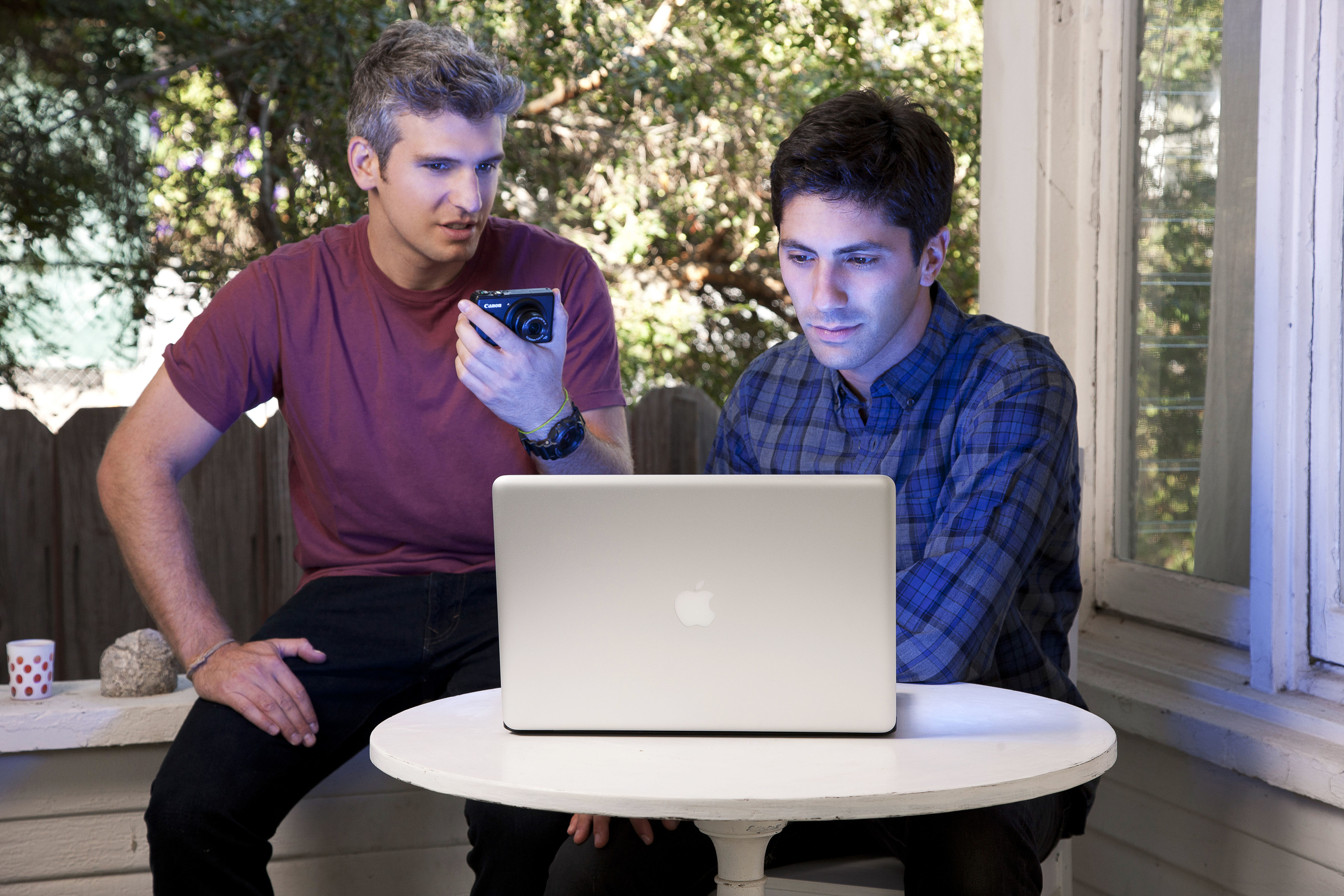 Two men, one with a smartphone, looking intently at a laptop screen on an outdoor patio