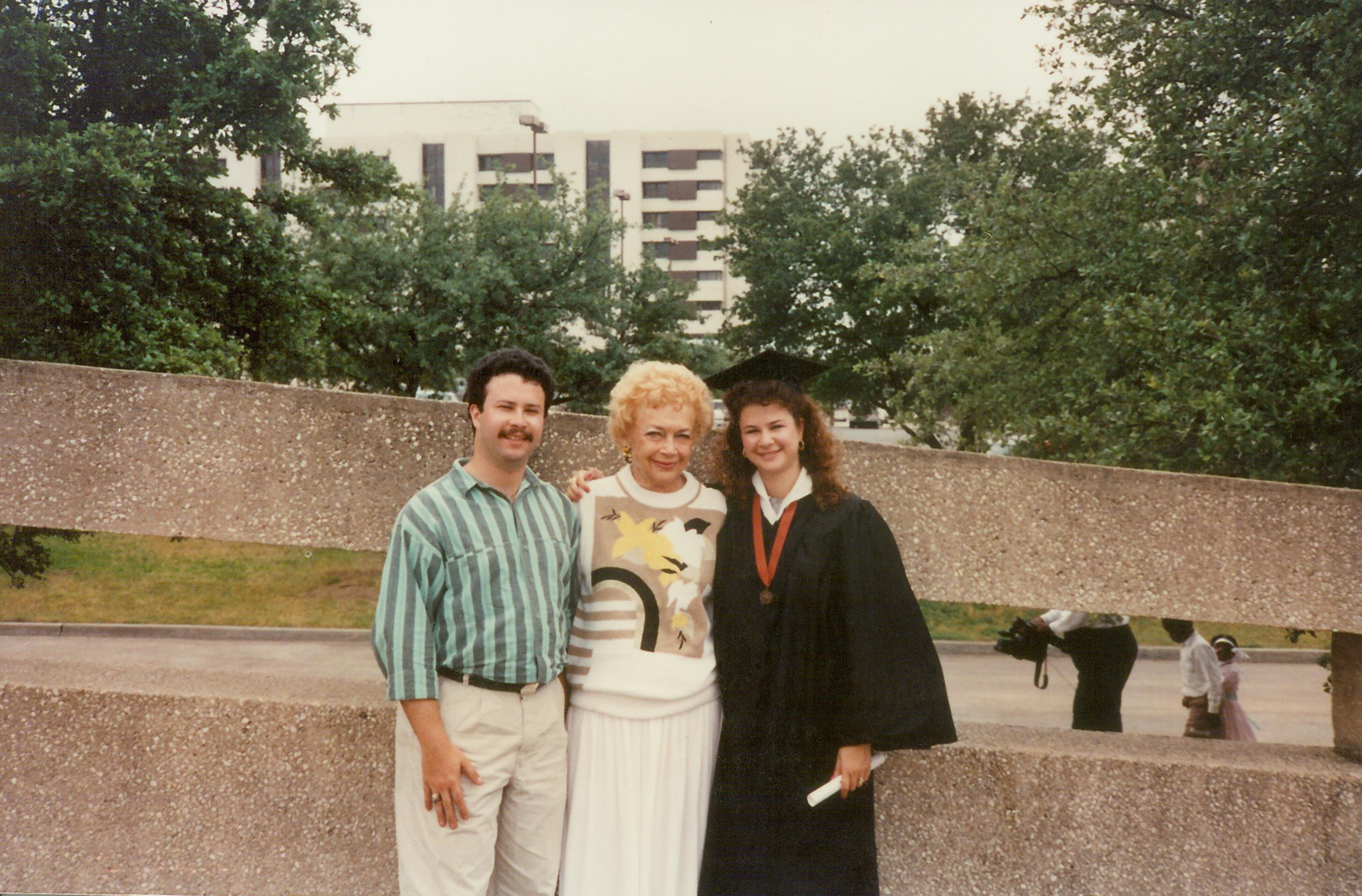 Three individuals posing outside, one in graduation attire with a diploma