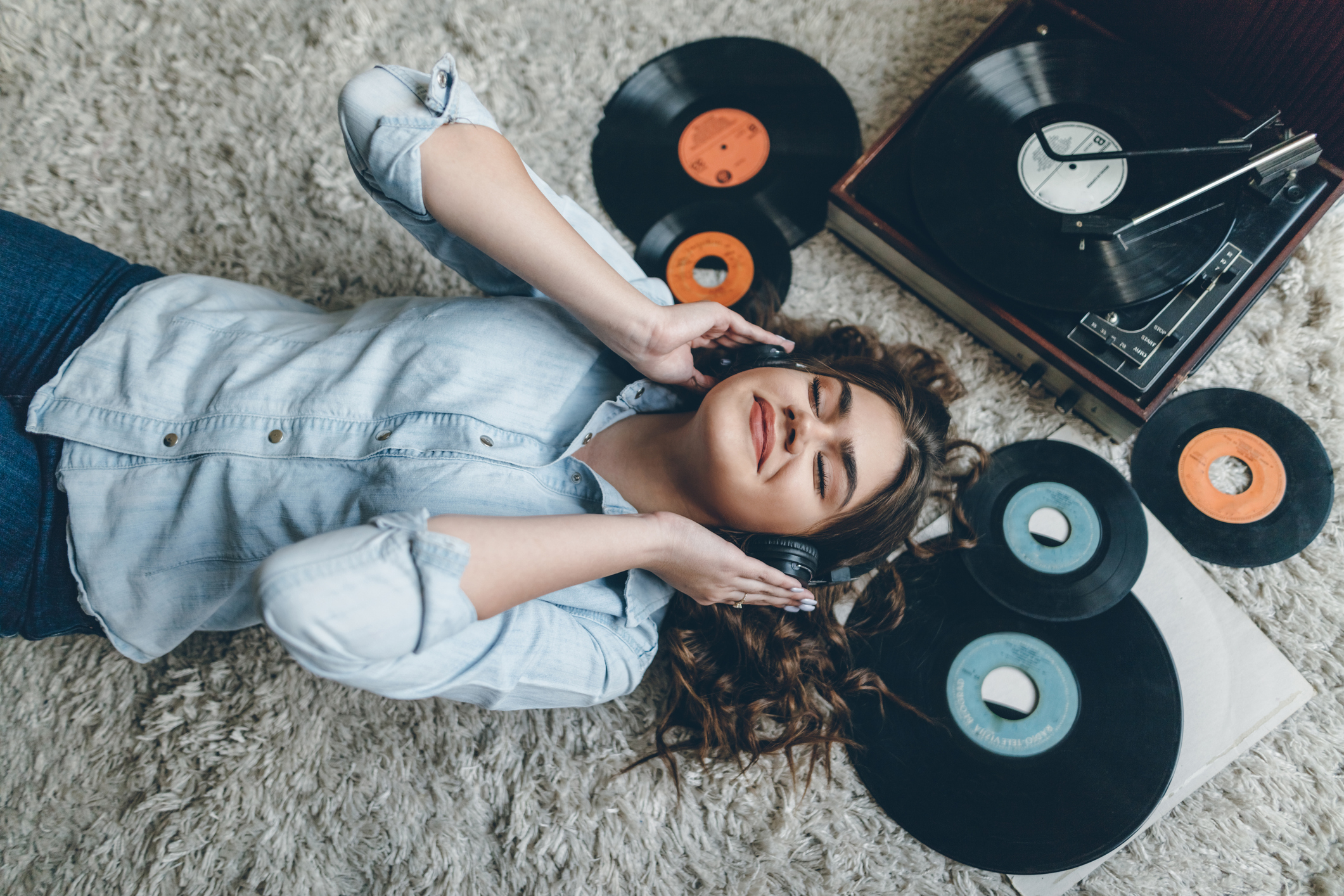 Woman lying on floor with headphones, surrounded by vinyl records and a turntable. She appears relaxed and happy