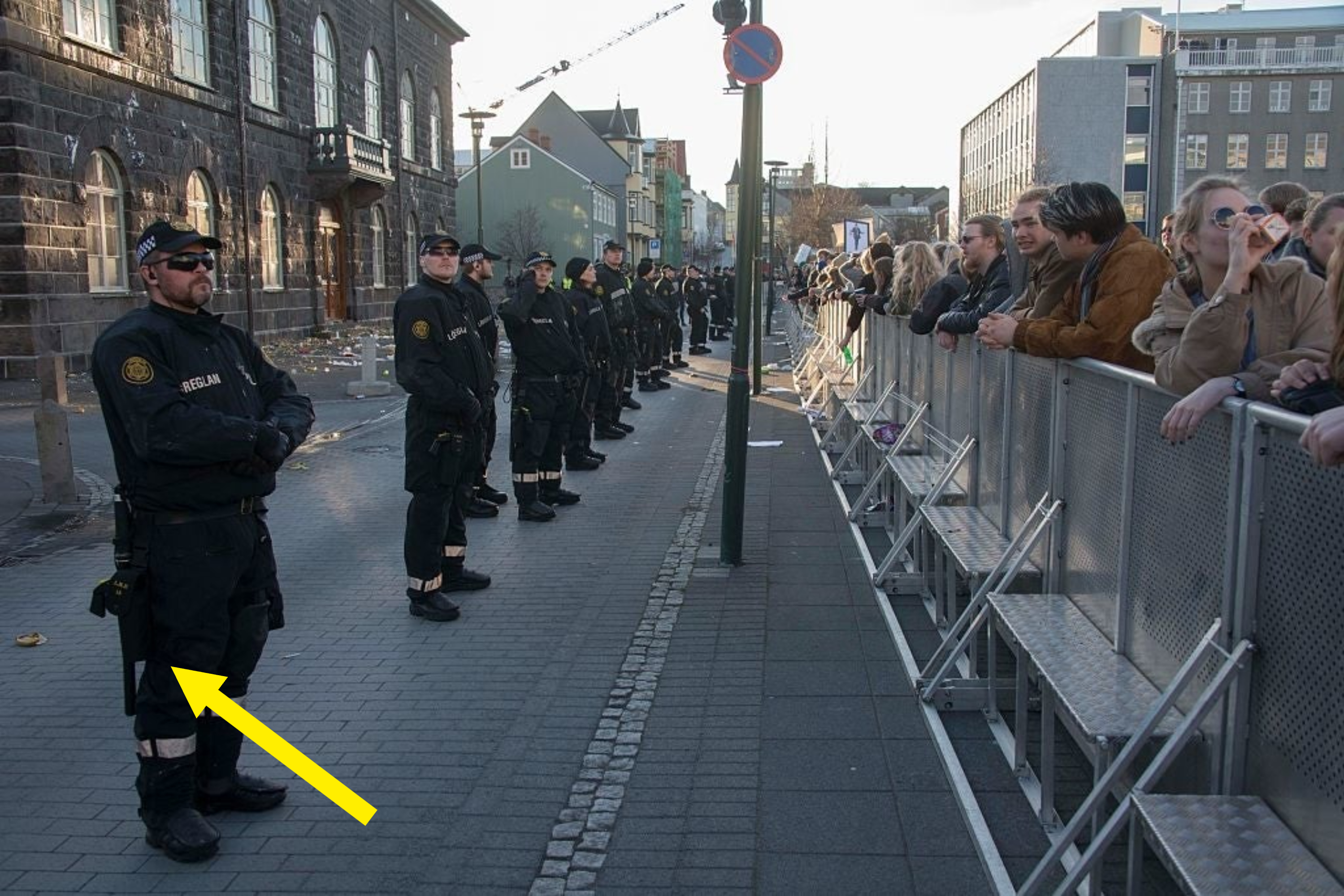 Police line the street with onlookers behind barriers, likely at a public event