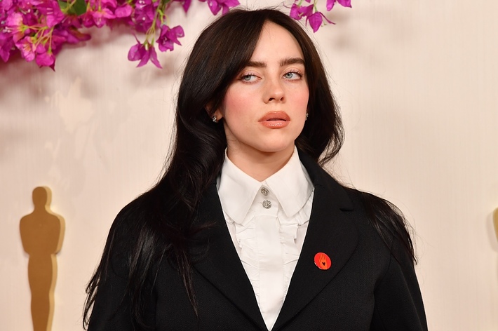 Billie Eilish at an event, wearing an oversized black suit with a white shirt and a red lapel pin