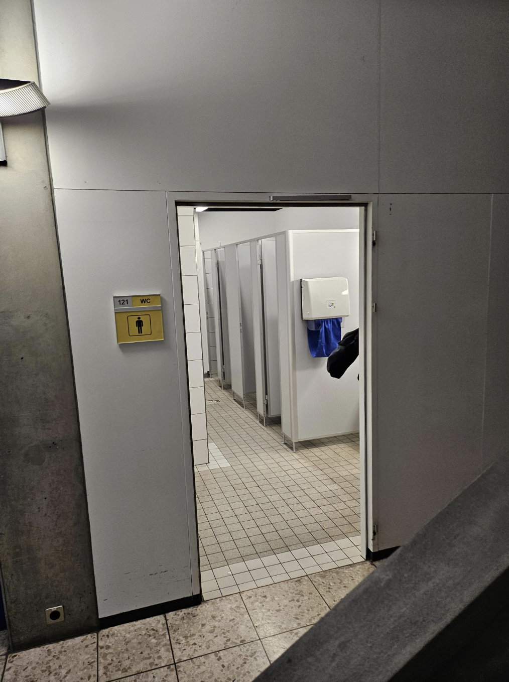 Public restroom interior with open stall doors and visible toilet paper dispenser; individual&#x27;s foot seen under one door
