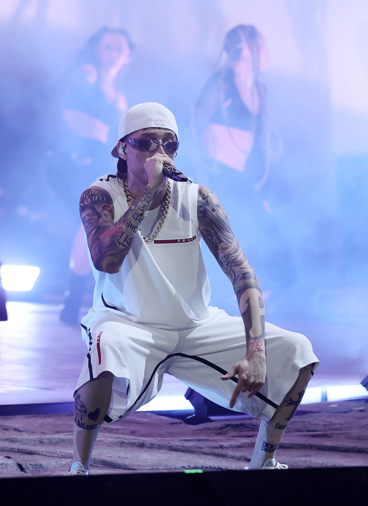 Peso Pluma performing on stage with a microphone, wearing a tank top, shorts, a hat, and sunglasses