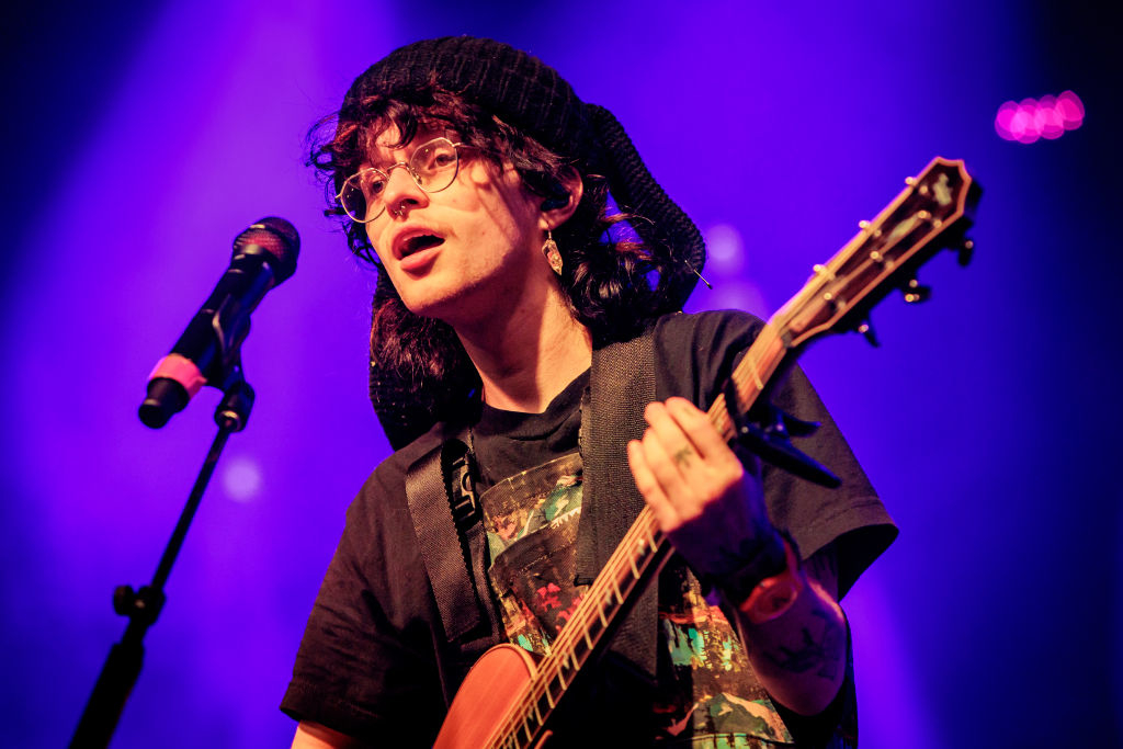 Cavetown with guitar onstage wearing a hat, graphic tee, and glasses