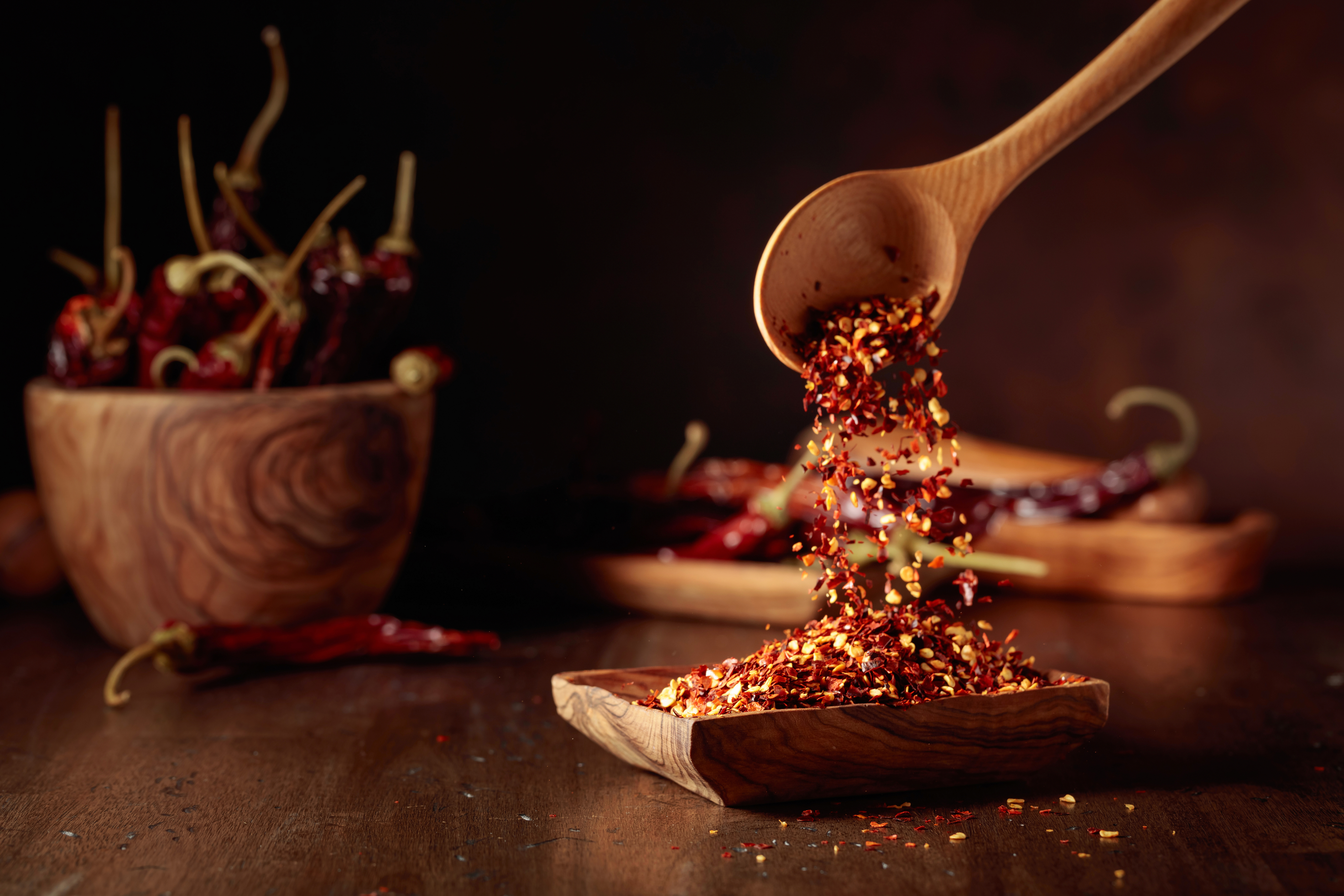Chili flakes being poured from a wooden spoon into a bowl with whole chilies in the background