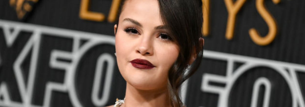 Selena Gomez at an event, wearing a jewel-necklace and dark lipstick, with a designed backdrop
