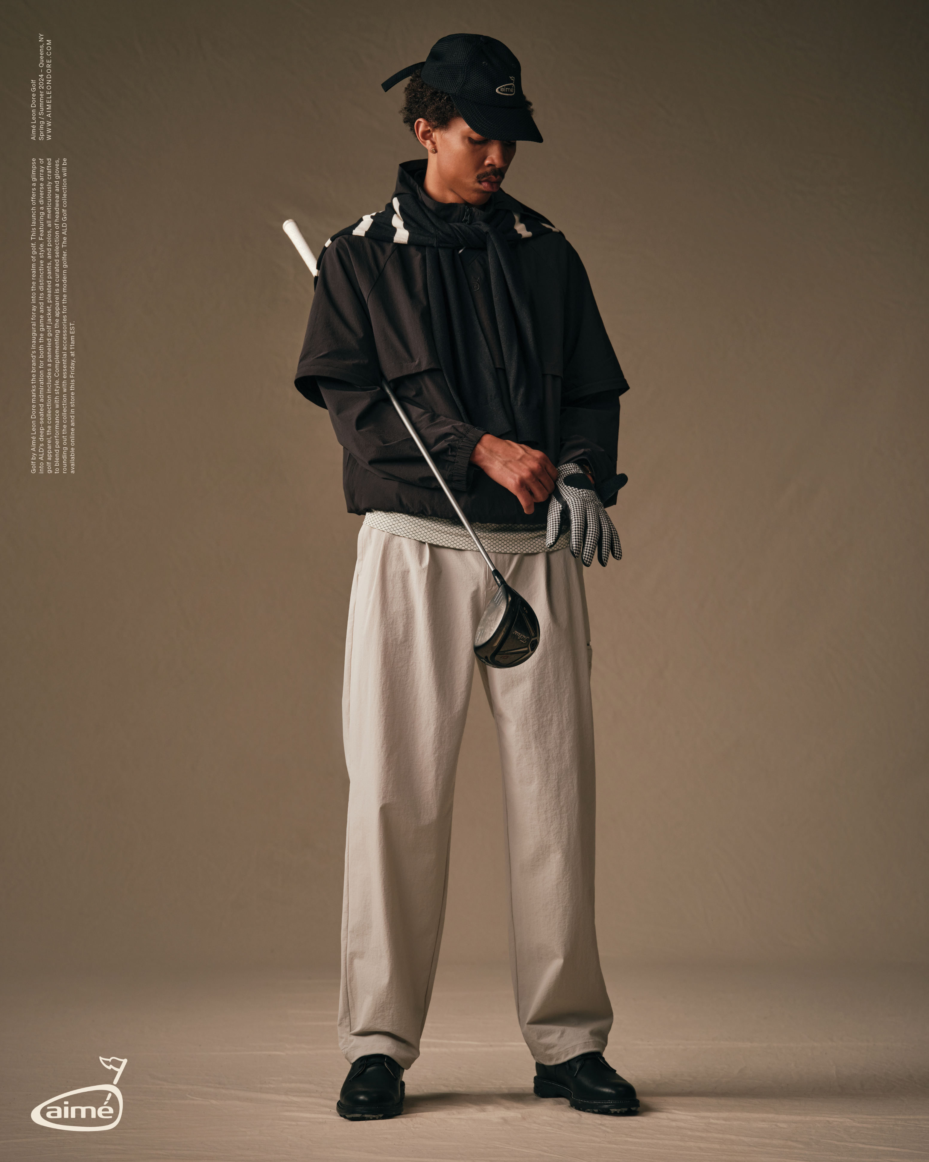 Person in a sporty ensemble with a jacket, cap, and draped neck accessory, holding a golf club, in a neutral-toned photoshoot