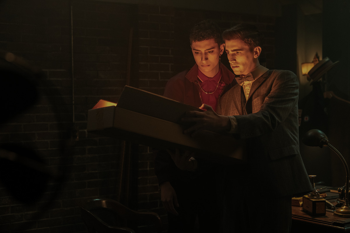 Dos actores, vestidos con trajes de época, mirando dentro de una caja con expresión intrigada en una escena de película o serie