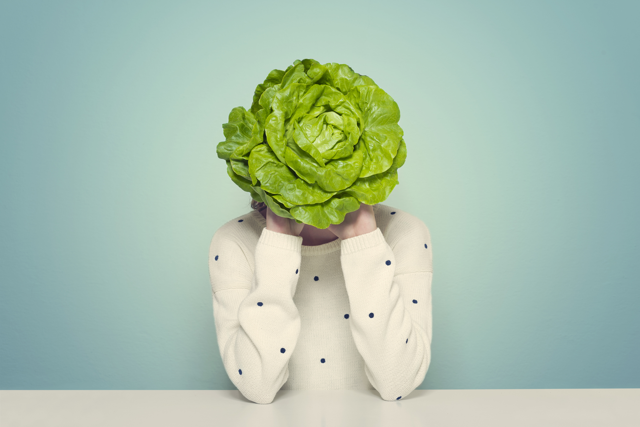 Person with a lettuce head obscuring their face, wearing a white sweater with dot patterns