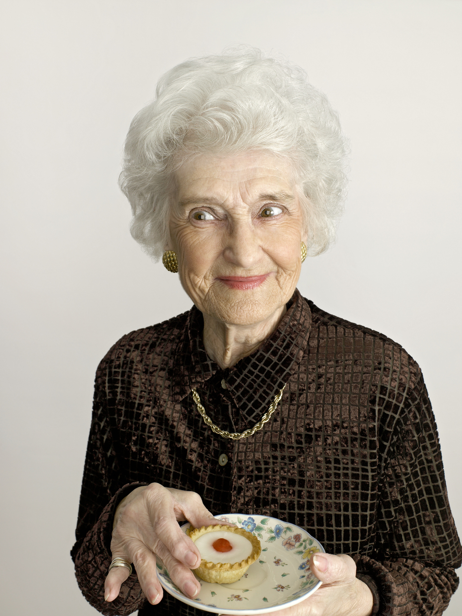 Elderly woman smiling, holding a plate with a pastry, dressed in a brown lace blouse and wearing earrings and a necklace